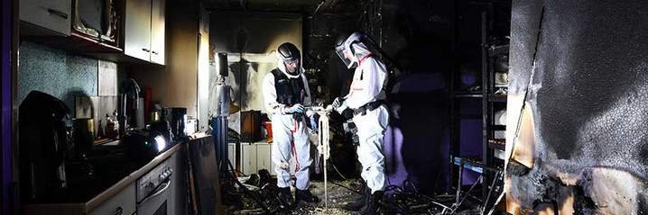 Zwei Brandermittler untersuchen einen Gegenstand in einem ausgebrannten Zimmer.
