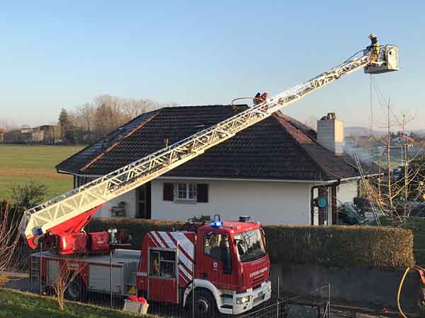 Ein Feuerwehrauto mit einer ausgefahrenen Leiter und einem darauf sich befindenden Feuerwehrmann befinden sich vor einem Haus, aus welchem Rauch aufsteigt.
