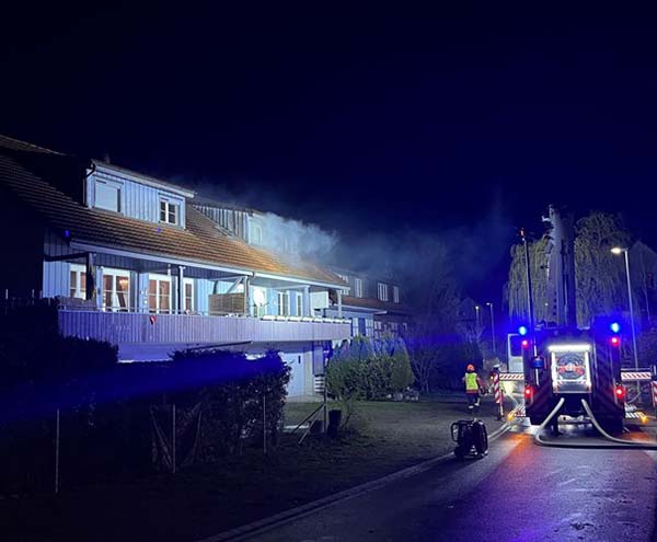 Die Feuerwehr mit einem Löschfahrzeug steht vor einem Haus, aus dem Rauch aufsteigt. 