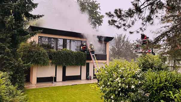 Aus einem Einfamilienhaus steigt Rauch auf. Feuerwehrleute auf einer Leiter sind das Feuer am Löschen.