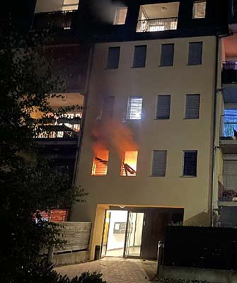 Aus zwei Fenstern eines Mehrfamilienhauses steigen Flammen und Rauch auf.